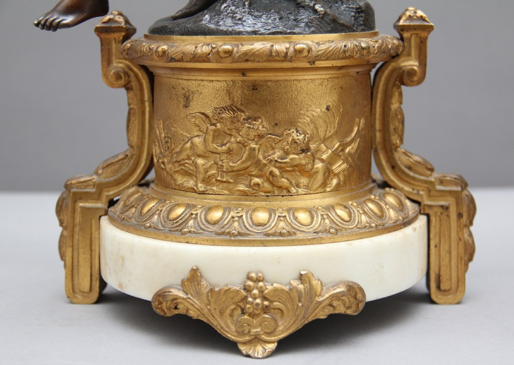 A pair of patinated dark bronze figurative statuettes depicting classical youths seated on rocky outcrops, set atop ornate gilded ormolu and white marble bases. View XII