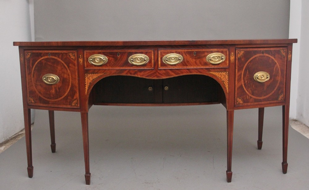 A 19th-century mahogany bowfront sideboard featuring marquetry inlay borders, brass oval handles, central arched drawer, and tapered legs.