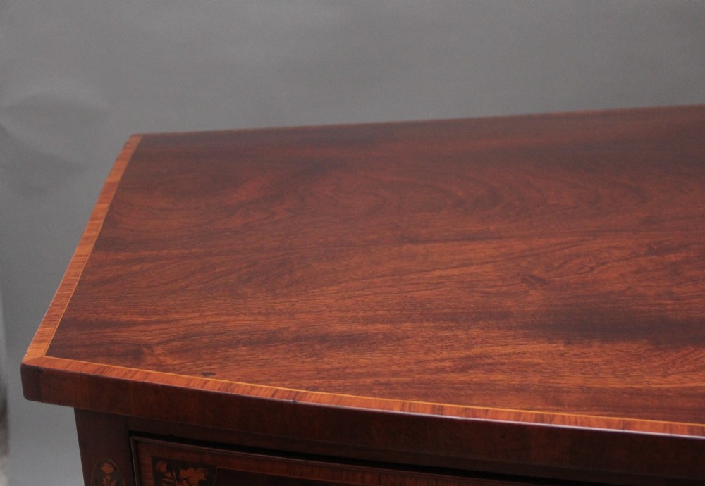 A 19th-century mahogany bowfront sideboard featuring marquetry inlay borders, brass oval handles, central arched drawer, and tapered legs. View X