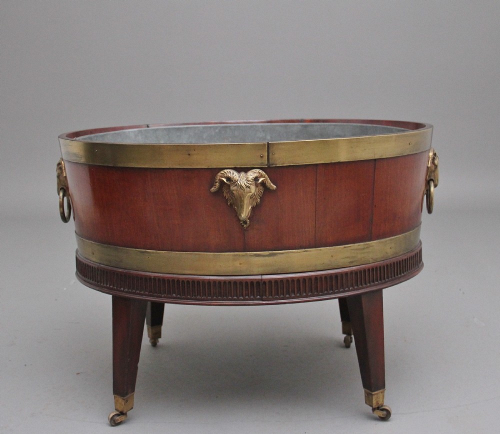 An oval early 19th-century mahogany wine cooler bound by thick brass bands, featuring a cast brass rams head mount, ring handles, and square tapered legs with brass castors.