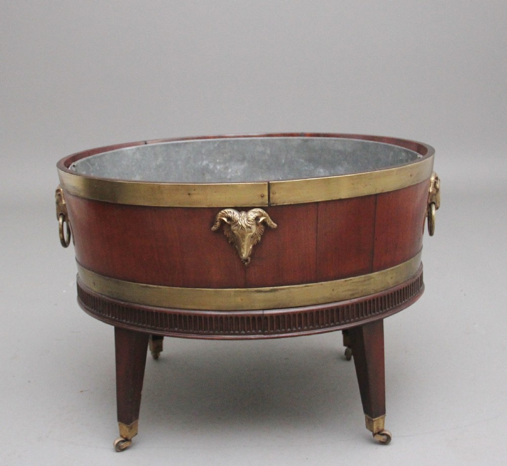 An oval early 19th-century mahogany wine cooler bound by thick brass bands, featuring a cast brass rams head mount, ring handles, and square tapered legs with brass castors. View II