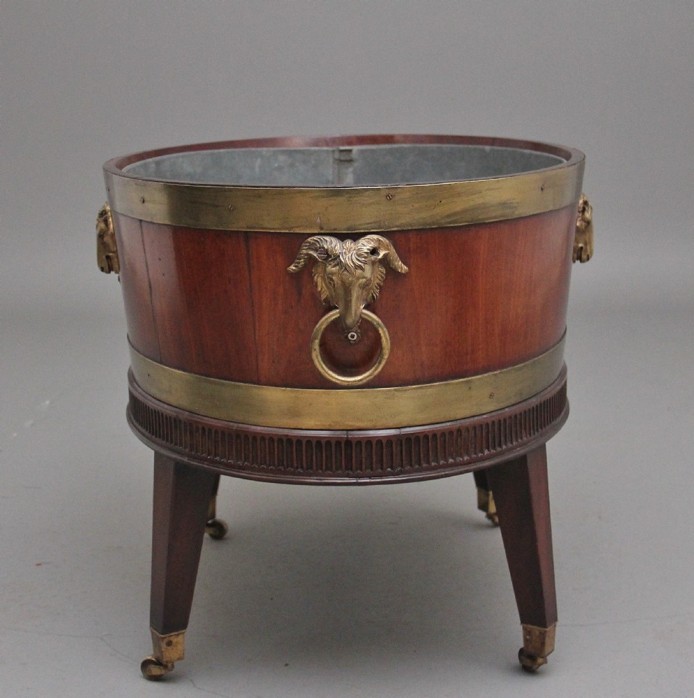 An oval early 19th-century mahogany wine cooler bound by thick brass bands, featuring a cast brass rams head mount, ring handles, and square tapered legs with brass castors. View IV