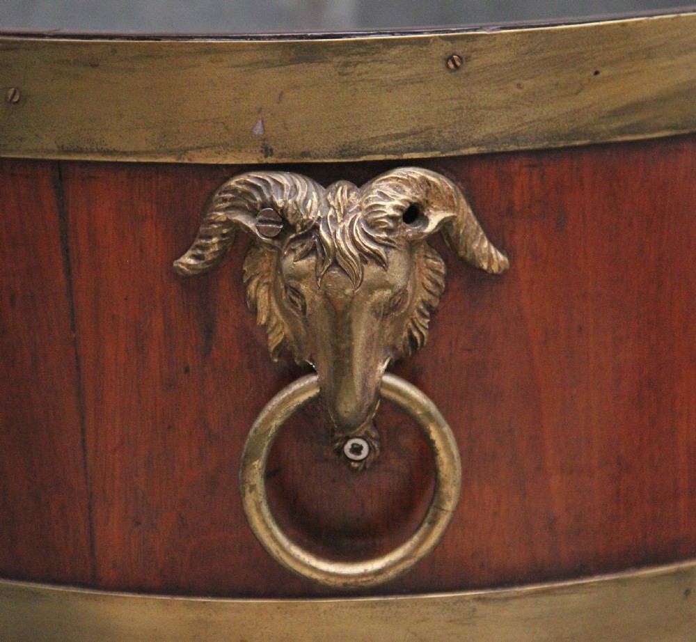An oval early 19th-century mahogany wine cooler bound by thick brass bands, featuring a cast brass rams head mount, ring handles, and square tapered legs with brass castors. View V