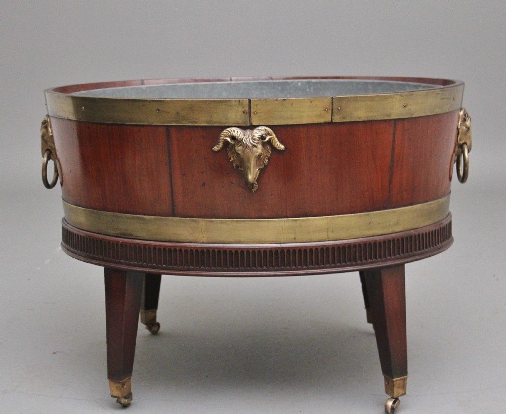 An oval early 19th-century mahogany wine cooler bound by thick brass bands, featuring a cast brass rams head mount, ring handles, and square tapered legs with brass castors. View VI