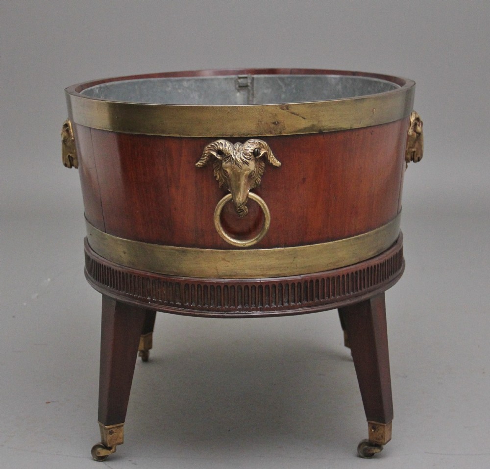 An oval early 19th-century mahogany wine cooler bound by thick brass bands, featuring a cast brass rams head mount, ring handles, and square tapered legs with brass castors. View VII