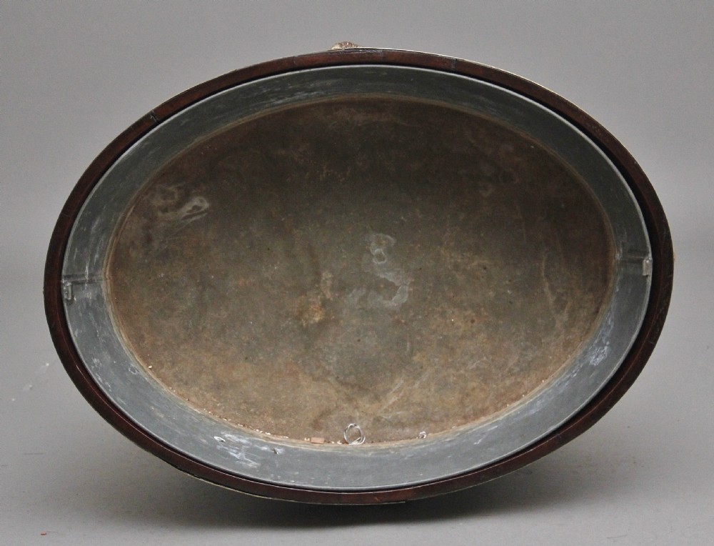 An oval early 19th-century mahogany wine cooler bound by thick brass bands, featuring a cast brass rams head mount, ring handles, and square tapered legs with brass castors. View VIII