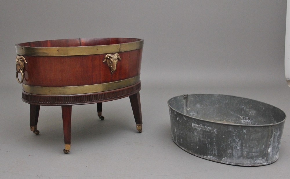 An oval early 19th-century mahogany wine cooler bound by thick brass bands, featuring a cast brass rams head mount, ring handles, and square tapered legs with brass castors. View IX