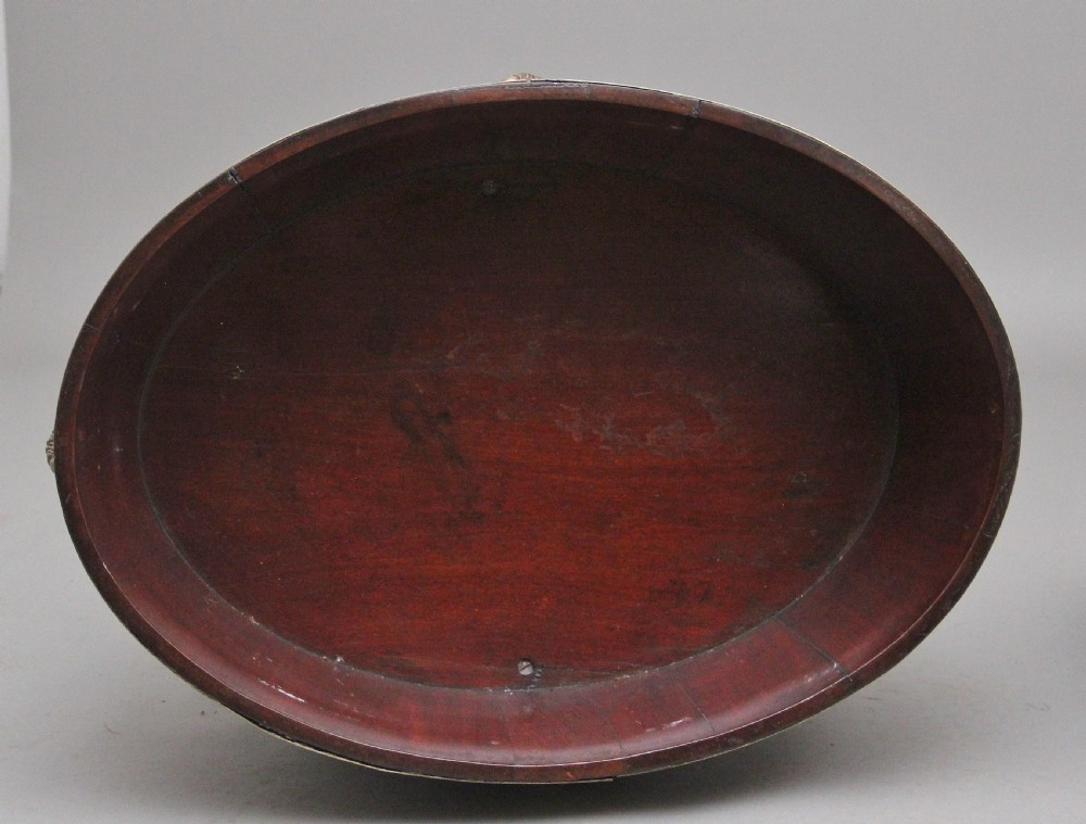 An oval early 19th-century mahogany wine cooler bound by thick brass bands, featuring a cast brass rams head mount, ring handles, and square tapered legs with brass castors. View X