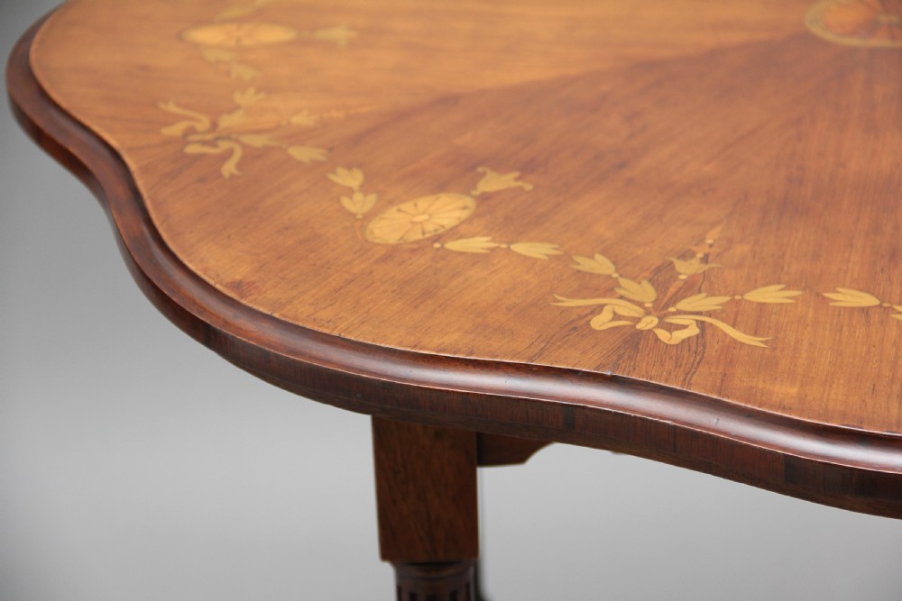 A 19th-century rosewood centre table with a serpentine-edged marquetry top, four fluted legs, an X-shaped stretcher, and brass castors. View X