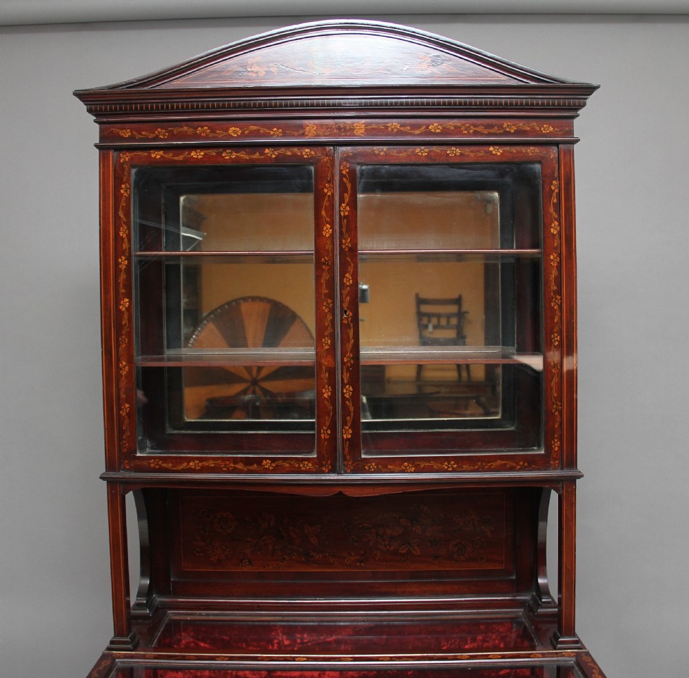 A 19th-century mahogany display cabinet featuring floral marquetry inlays, a glazed upper section with glass shelves, and a bow-front lower cabinet on tapered legs. View VII