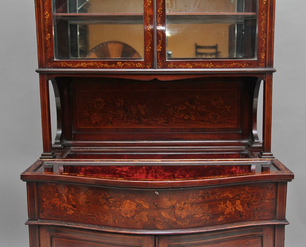 A 19th-century mahogany display cabinet featuring floral marquetry inlays, a glazed upper section with glass shelves, and a bow-front lower cabinet on tapered legs. View VIII