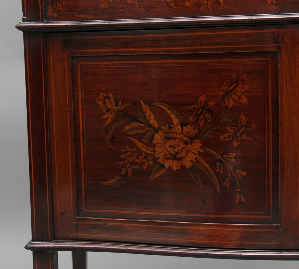 A 19th-century mahogany display cabinet featuring floral marquetry inlays, a glazed upper section with glass shelves, and a bow-front lower cabinet on tapered legs. View XVI