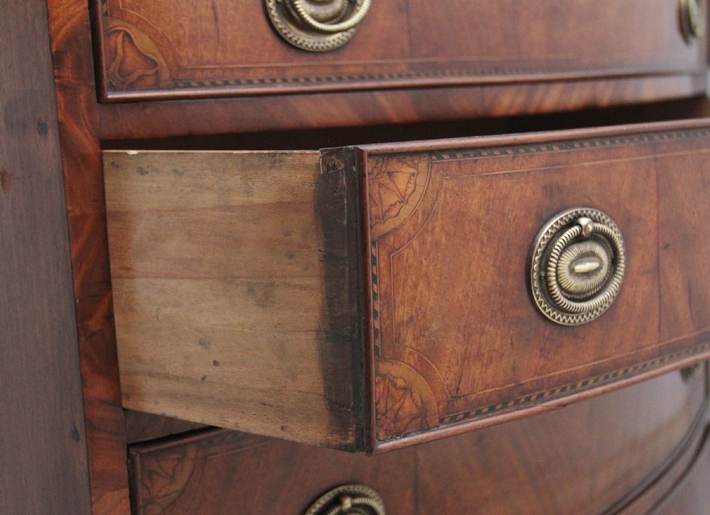 An 18th-century bowfront mahogany chest featuring four graduated drawers with oval brass ring handles, decorative inlay, and splayed bracket feet. View VI