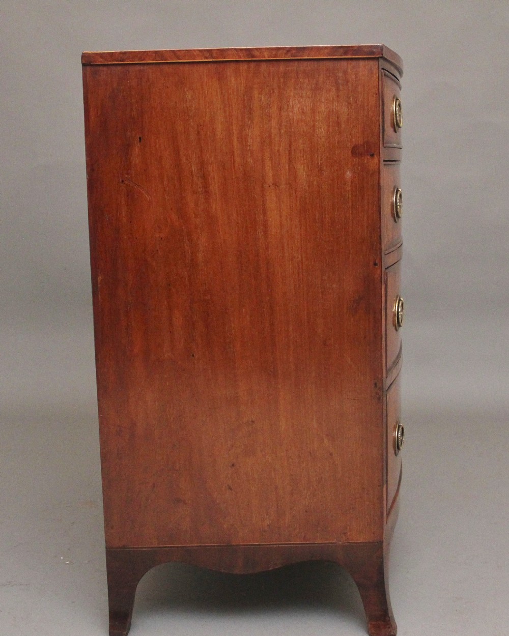 An 18th-century bowfront mahogany chest featuring four graduated drawers with oval brass ring handles, decorative inlay, and splayed bracket feet. View VII