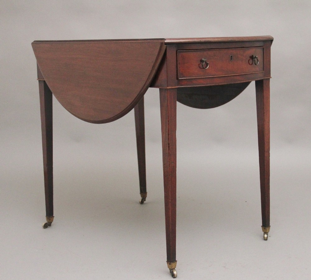 An 18th-century mahogany Pembroke table featuring drop-down curved side leaves, a front drawer with ring pull handles, and square tapered legs on brass castors.