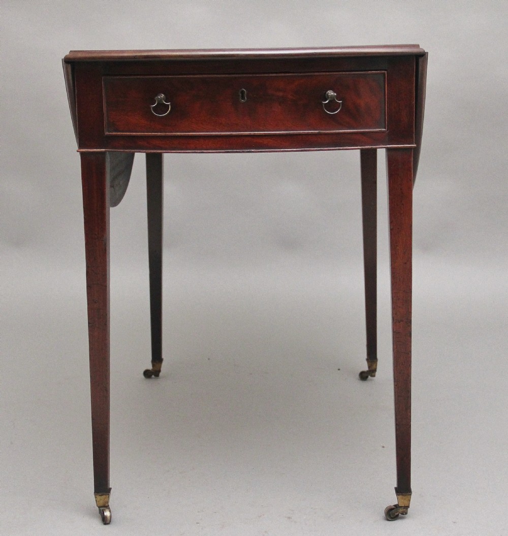 An 18th-century mahogany Pembroke table featuring drop-down curved side leaves, a front drawer with ring pull handles, and square tapered legs on brass castors. View IV