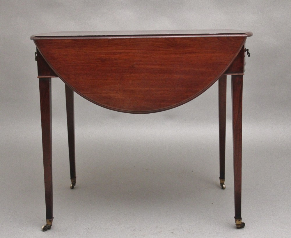 An 18th-century mahogany Pembroke table featuring drop-down curved side leaves, a front drawer with ring pull handles, and square tapered legs on brass castors. View VI