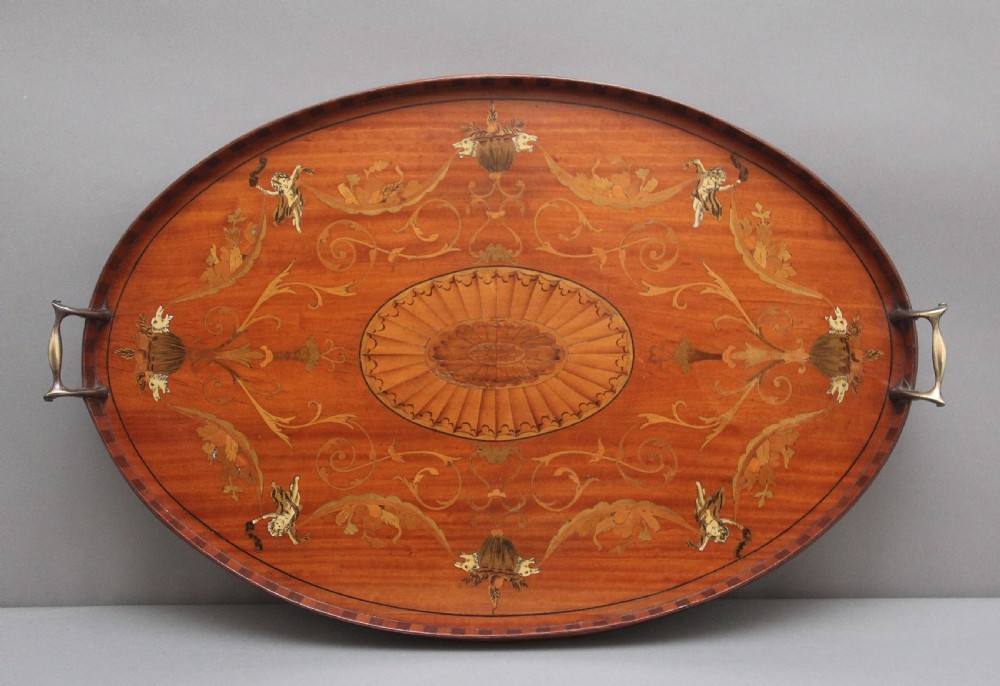 An oval 19th-century satinwood tray featuring detailed marquetry inlays of cherubs, foliate scrolls, a central sunburst medallion, and brass handles at each end.