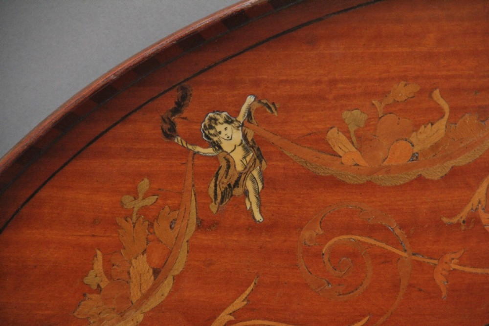 An oval 19th-century satinwood tray featuring detailed marquetry inlays of cherubs, foliate scrolls, a central sunburst medallion, and brass handles at each end. View V