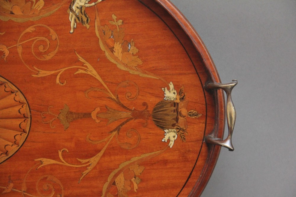 An oval 19th-century satinwood tray featuring detailed marquetry inlays of cherubs, foliate scrolls, a central sunburst medallion, and brass handles at each end. View VII