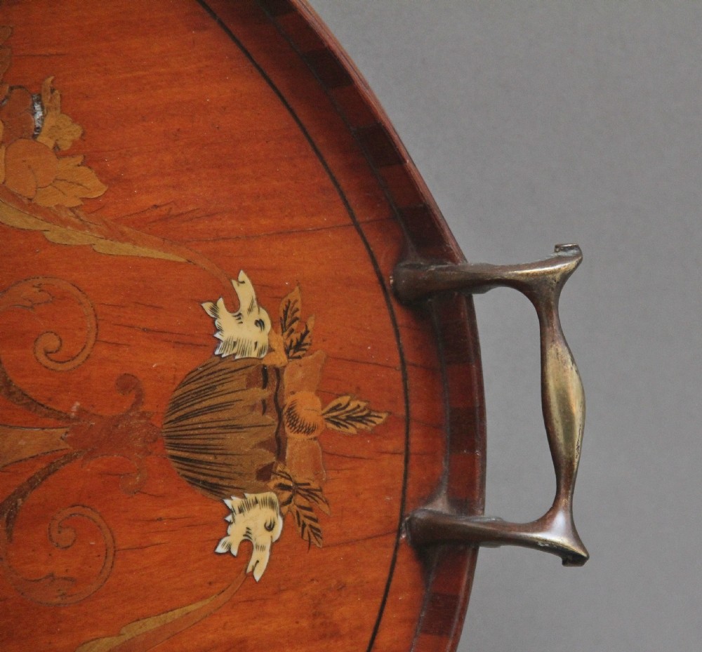 An oval 19th-century satinwood tray featuring detailed marquetry inlays of cherubs, foliate scrolls, a central sunburst medallion, and brass handles at each end. View IX