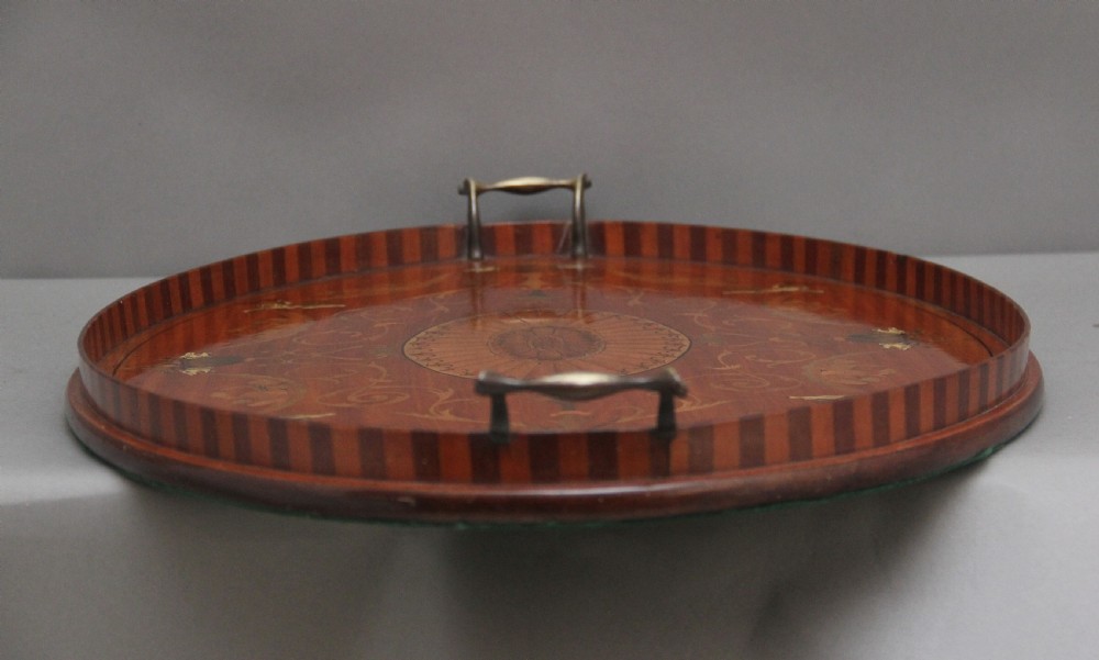 An oval 19th-century satinwood tray featuring detailed marquetry inlays of cherubs, foliate scrolls, a central sunburst medallion, and brass handles at each end. View X