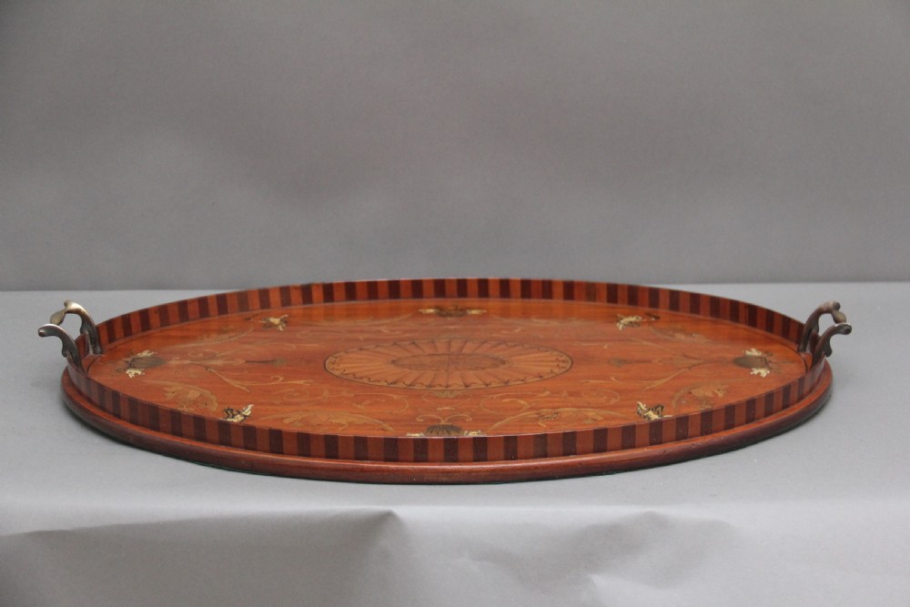 An oval 19th-century satinwood tray featuring detailed marquetry inlays of cherubs, foliate scrolls, a central sunburst medallion, and brass handles at each end. View XI