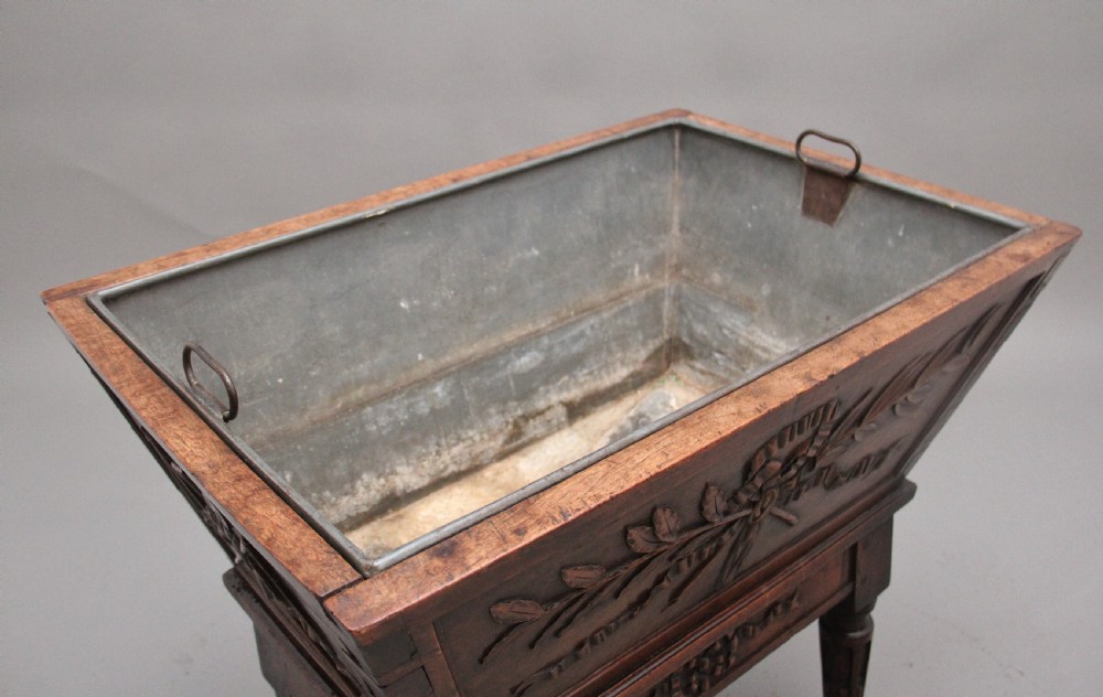 An 18th-century French carved walnut wine cooler featuring a zinc-lined tapered basin set on a stand with fluted legs and ribbon relief carvings. View V