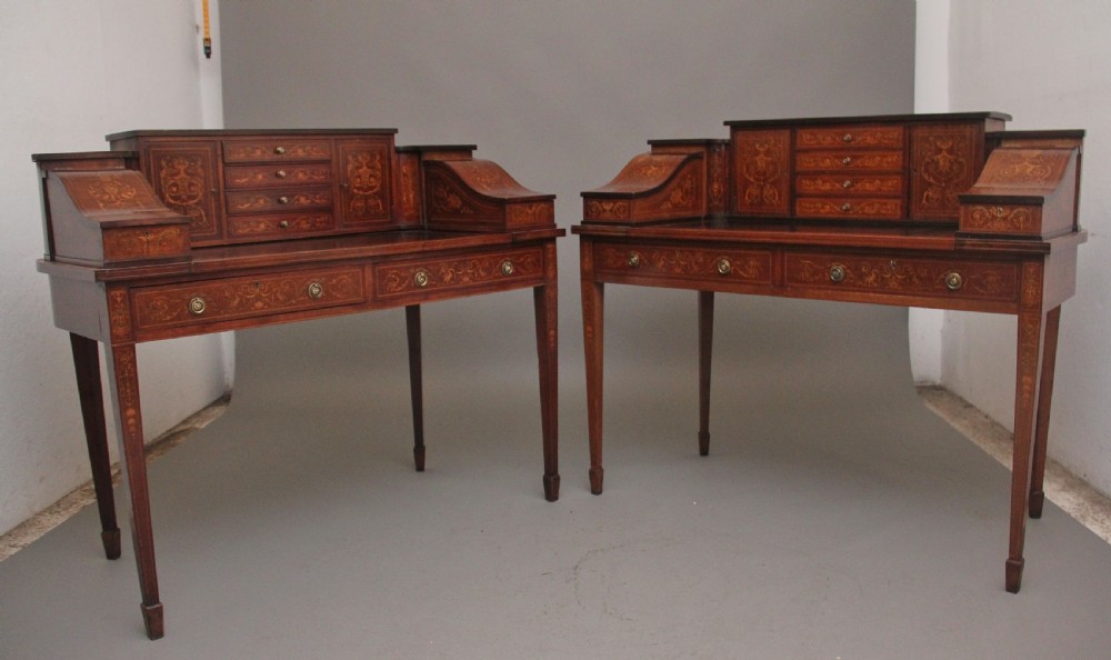 A pair of early 20th-century mahogany Carlton House desks with detailed satinwood marquetry inlay, stepped gallery drawers, and tapered legs.