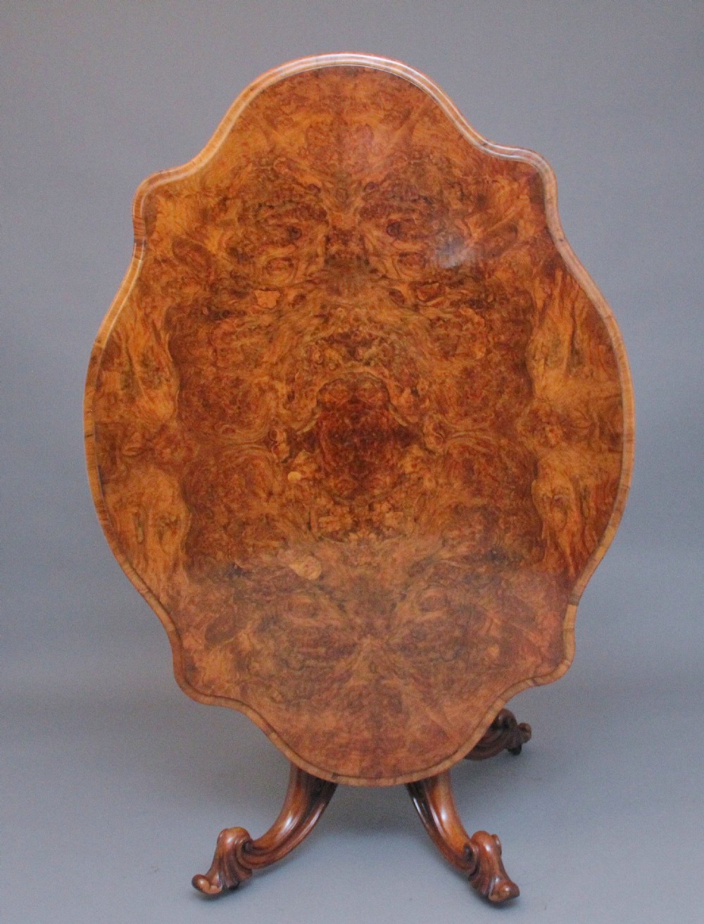 A 19th-century Victorian burr walnut centre table with a shaped, highly figured tilt-top top, raised on a carved pedestal with four cabriole legs on brass castors. View II