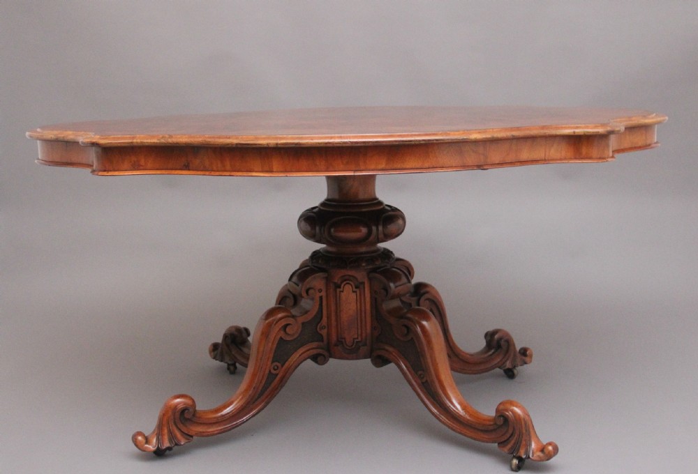 A 19th-century Victorian burr walnut centre table with a shaped, highly figured tilt-top top, raised on a carved pedestal with four cabriole legs on brass castors. View VI
