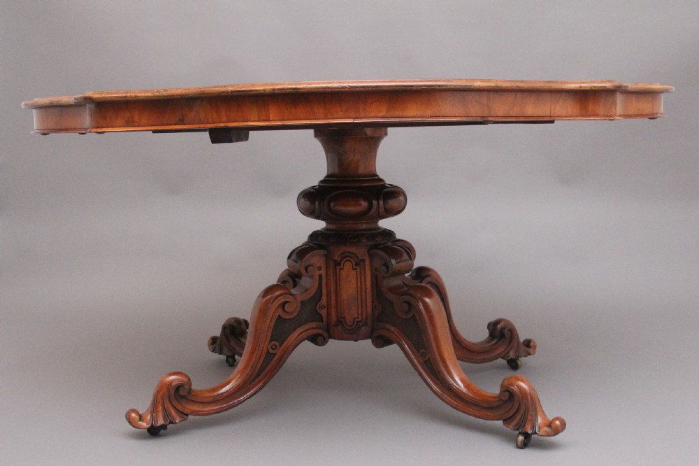 A 19th-century Victorian burr walnut centre table with a shaped, highly figured tilt-top top, raised on a carved pedestal with four cabriole legs on brass castors. View VII