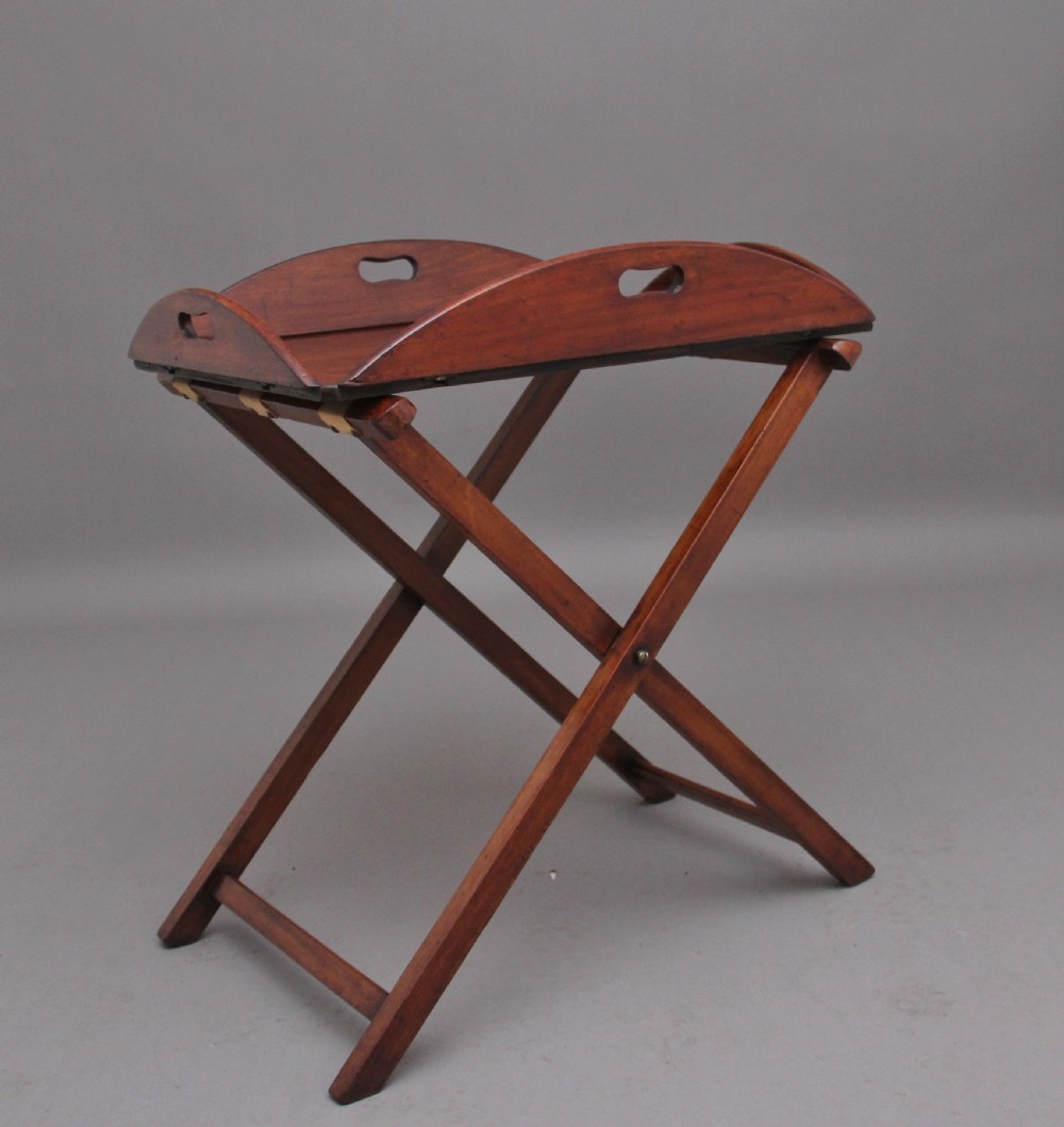 A 19th-century mahogany butler tray featuring shaped gallery sides with cutout handles resting upon a collapsible X-frame stand with webbing straps.