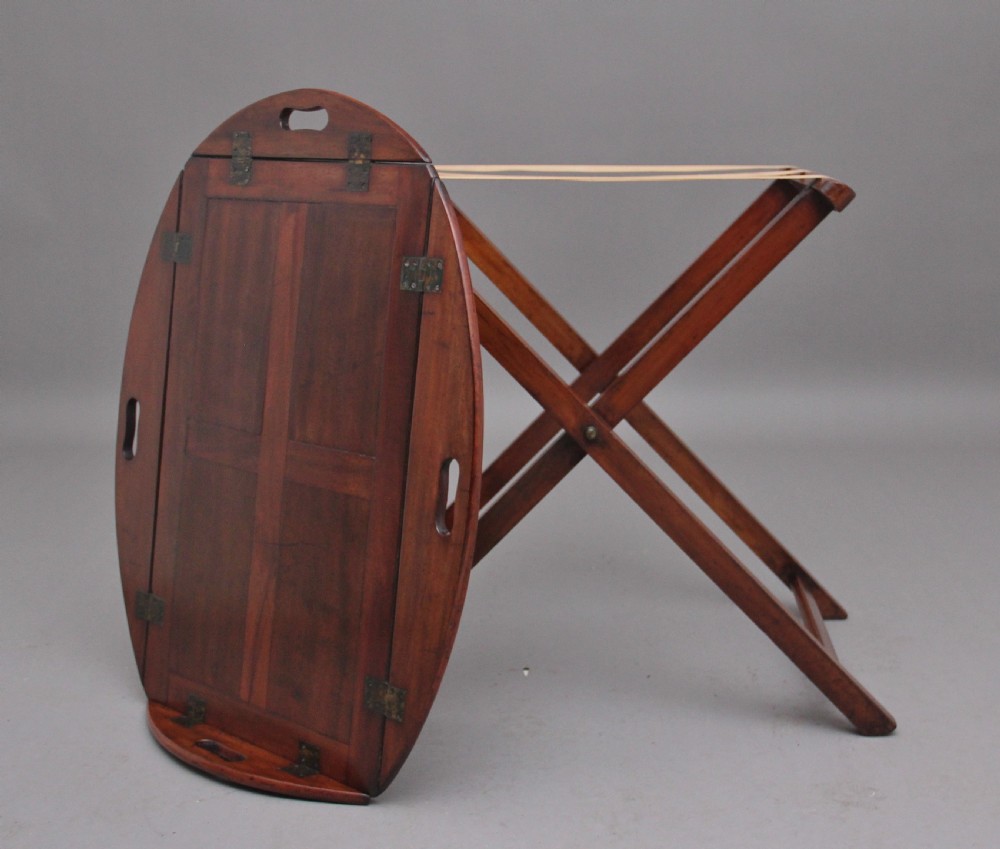 A 19th-century mahogany butler tray featuring shaped gallery sides with cutout handles resting upon a collapsible X-frame stand with webbing straps. View X