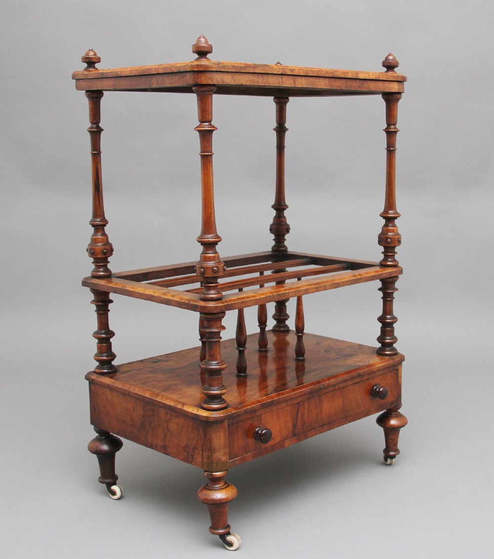 A Victorian burr walnut Canterbury featuring a flat top surface, open slatted partitions, a single base drawer, turned supports, and ceramic castors.
