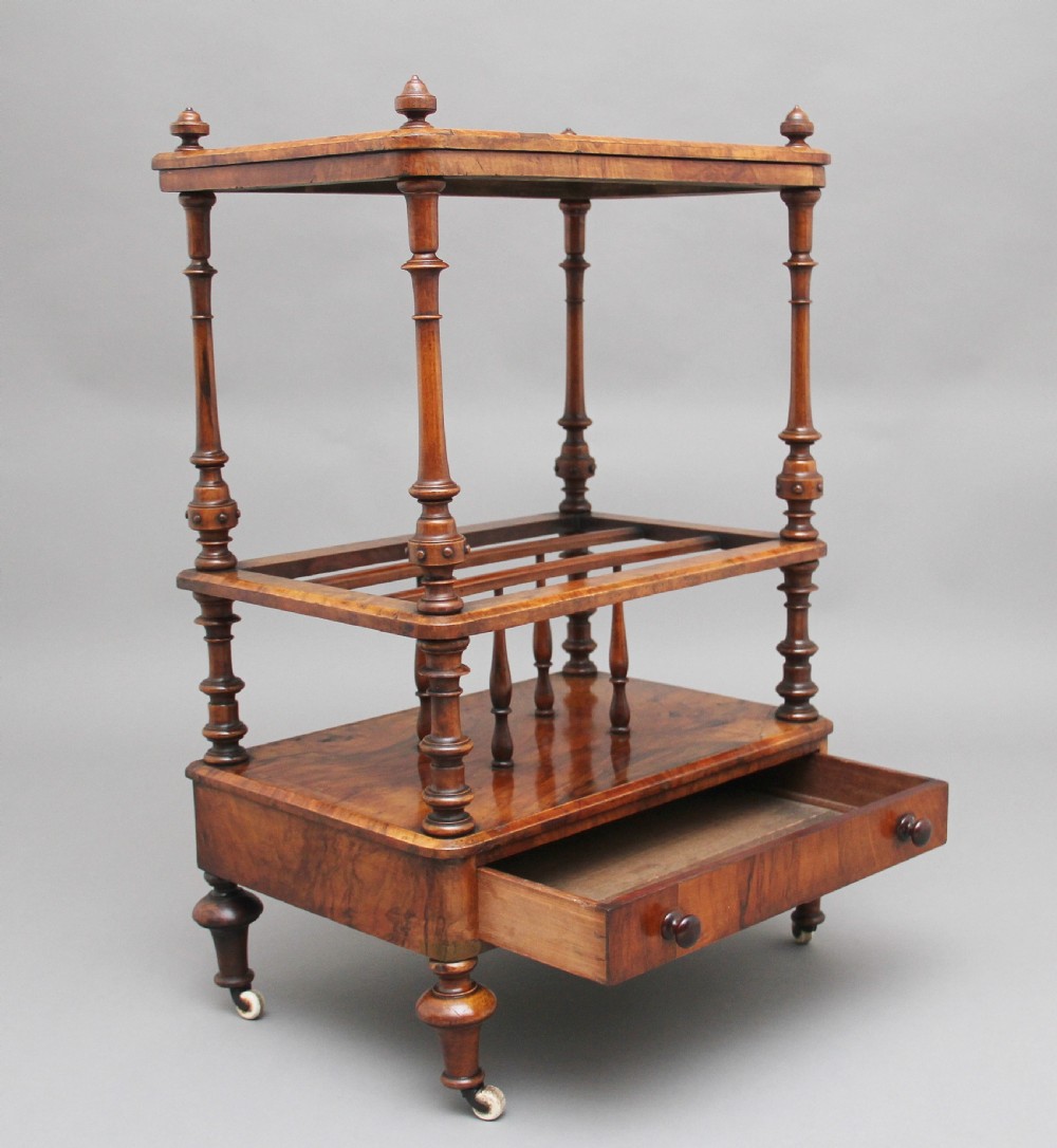 A Victorian burr walnut Canterbury featuring a flat top surface, open slatted partitions, a single base drawer, turned supports, and ceramic castors. View II