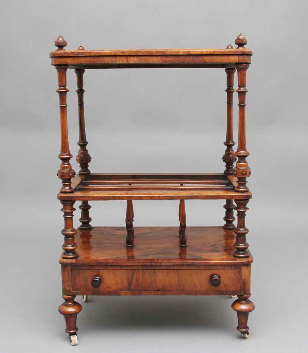 A Victorian burr walnut Canterbury featuring a flat top surface, open slatted partitions, a single base drawer, turned supports, and ceramic castors. View III