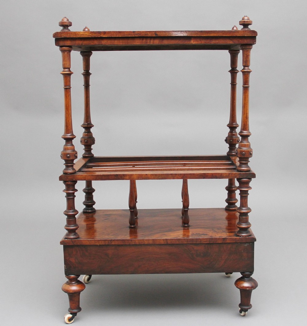A Victorian burr walnut Canterbury featuring a flat top surface, open slatted partitions, a single base drawer, turned supports, and ceramic castors. View VI