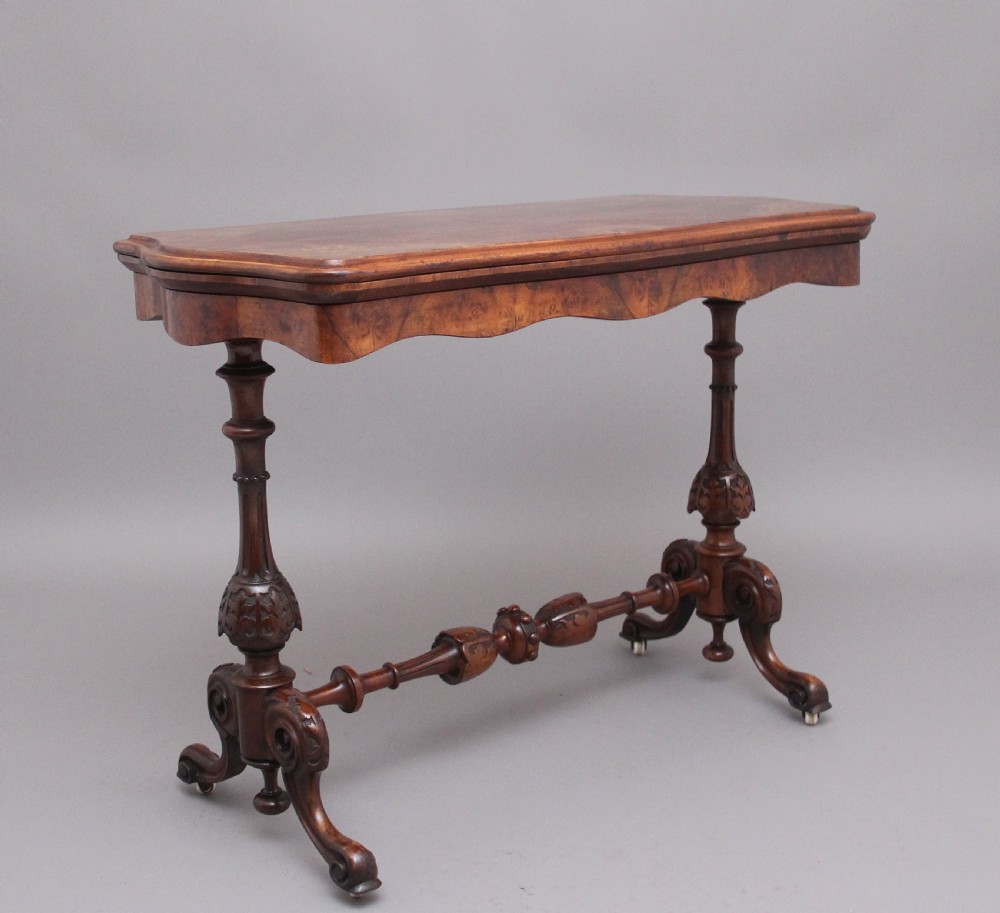 A Victorian burr walnut fold-over card table featuring a serpentine top, carved turned supports, and a stretcher base on castors.