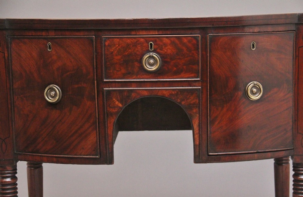 An early 19th-century bowfront mahogany sideboard with turned legs, brass ring drop handles, and central arch drawer. View V