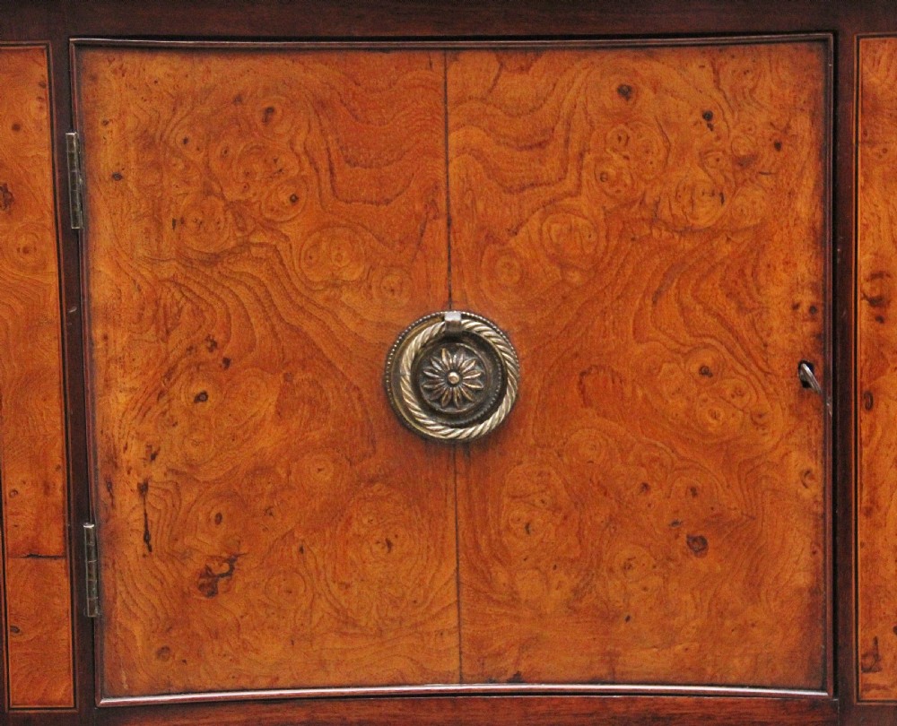 A nineteenth-century mahogany sideboard featuring burr elm veneer panels, two side cupboards, a central drawer above an arched apron, and four tapered square legs with brass ring handles. View V