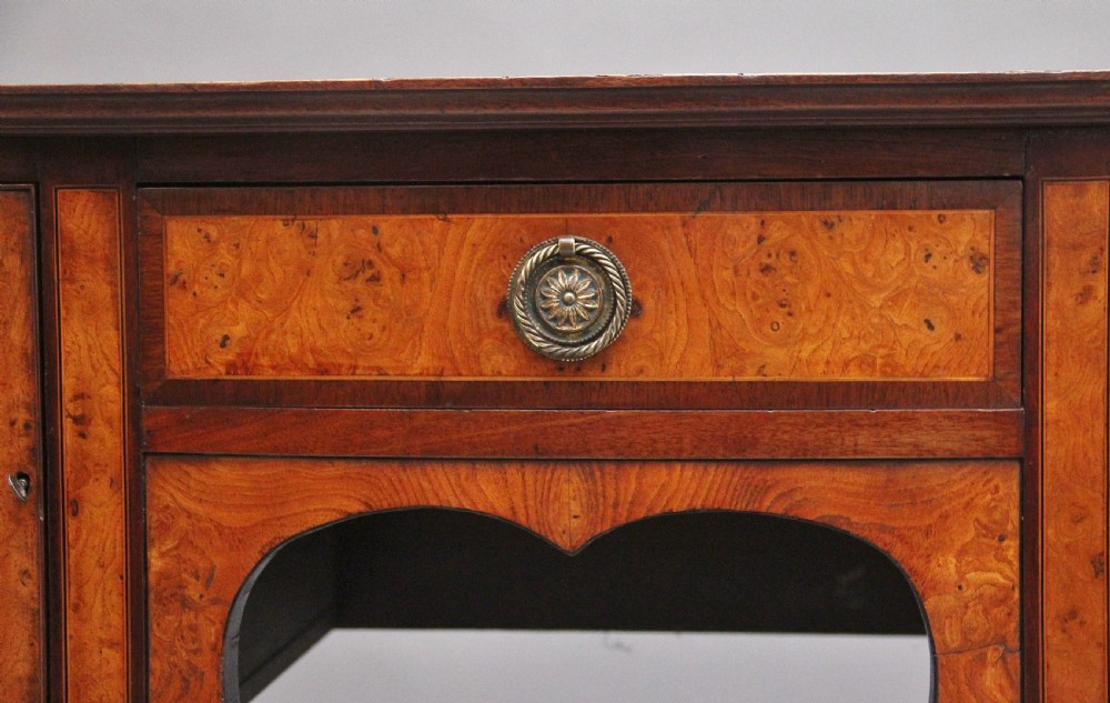 A nineteenth-century mahogany sideboard featuring burr elm veneer panels, two side cupboards, a central drawer above an arched apron, and four tapered square legs with brass ring handles. View VI