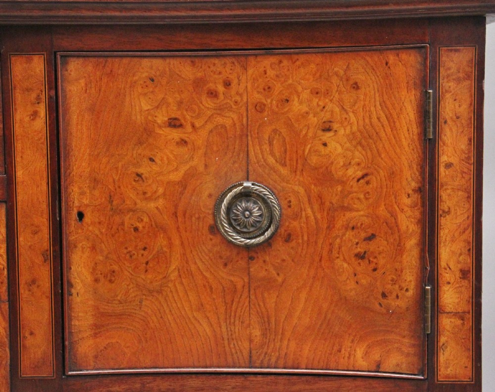 A nineteenth-century mahogany sideboard featuring burr elm veneer panels, two side cupboards, a central drawer above an arched apron, and four tapered square legs with brass ring handles. View VII
