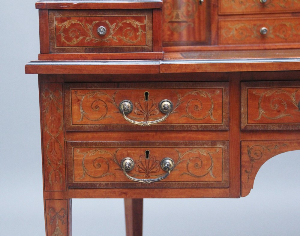 A 19th-century satinwood Carlton House writing desk featuring decorative marquetry, small drawers on a U-shaped superstructure, and tapered square legs with brass castors. View XVI