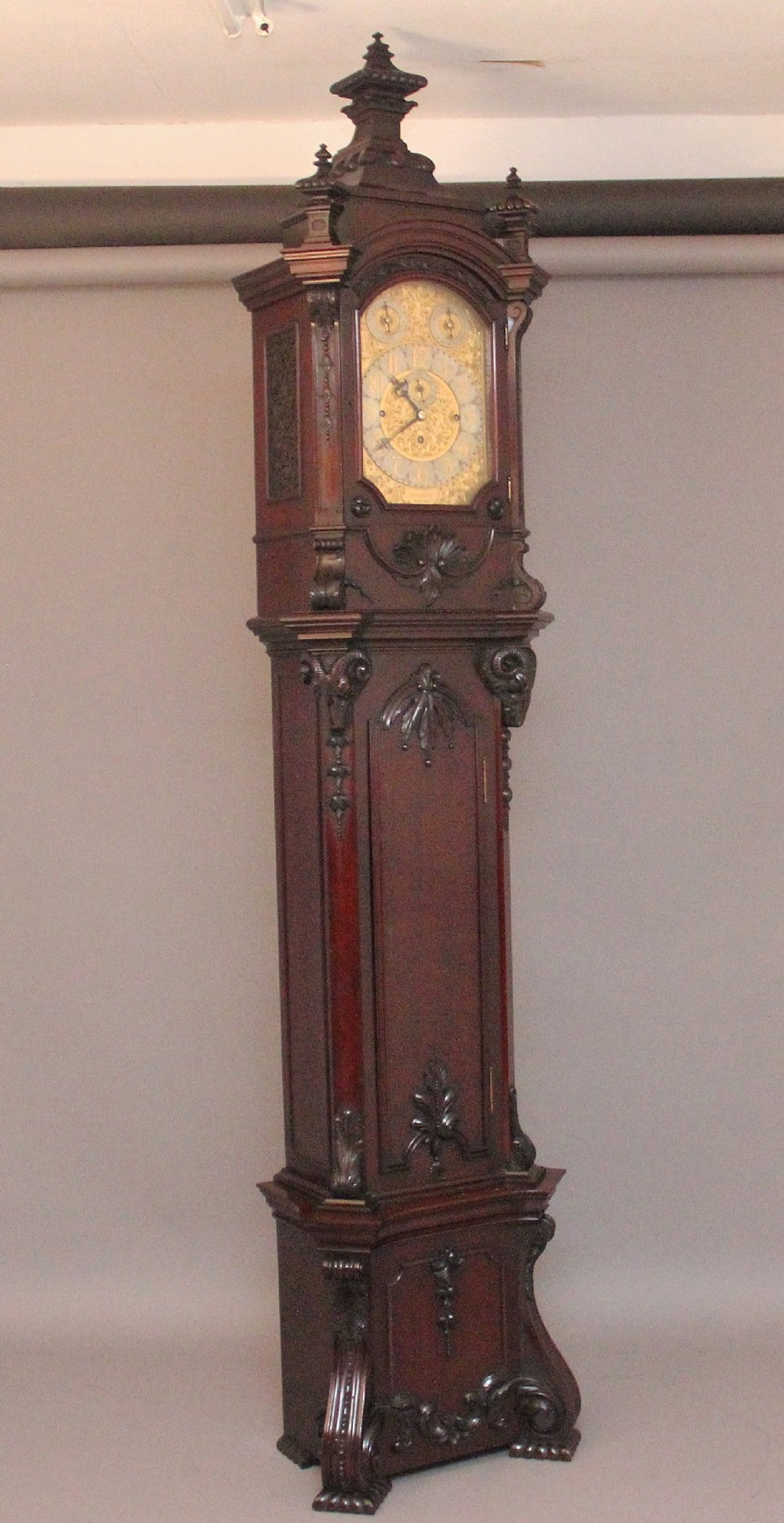 An early 20th-century carved dark mahogany longcase clock featuring an ornate pediment, carved trunk door, and etched brass dial.