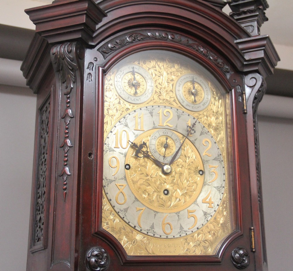 An early 20th-century carved dark mahogany longcase clock featuring an ornate pediment, carved trunk door, and etched brass dial. View VI