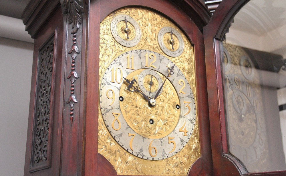 An early 20th-century carved dark mahogany longcase clock featuring an ornate pediment, carved trunk door, and etched brass dial. View VII