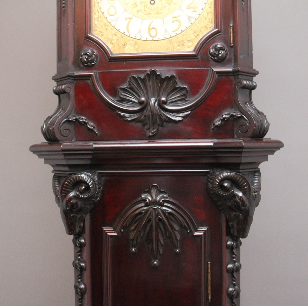 An early 20th-century carved dark mahogany longcase clock featuring an ornate pediment, carved trunk door, and etched brass dial. View IX