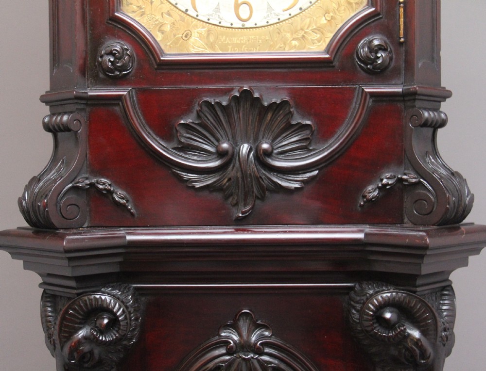 An early 20th-century carved dark mahogany longcase clock featuring an ornate pediment, carved trunk door, and etched brass dial. View X