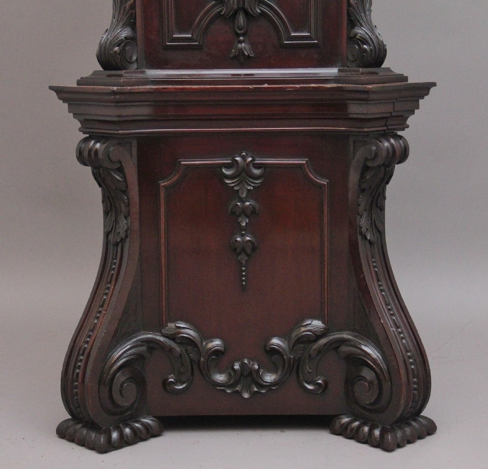 An early 20th-century carved dark mahogany longcase clock featuring an ornate pediment, carved trunk door, and etched brass dial. View XIV